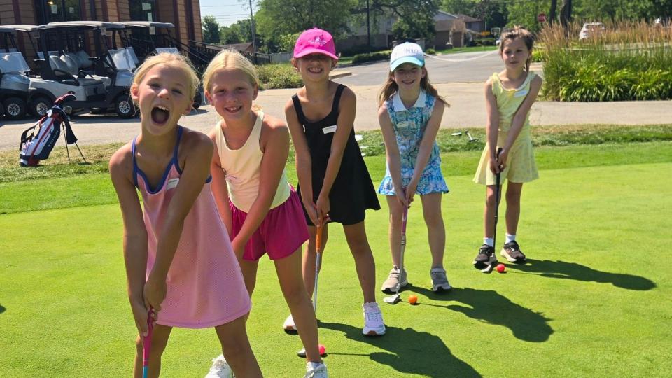 Five children in golf attire stand on a putting green, each holding a putter and smiling at the camera.